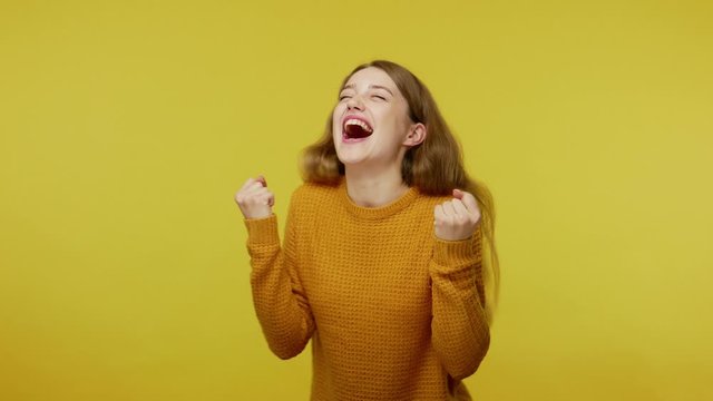 Excited champion. Enthusiastic overjoyed beautiful girl in pullover rejoicing victory, celebrating success with raised fists and dance, shouting yes. indoor studio shot isolated on yellow background