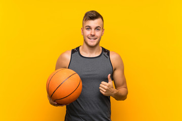 Young handsome blonde man holding a basket ball over isolated yellow background with thumbs up...