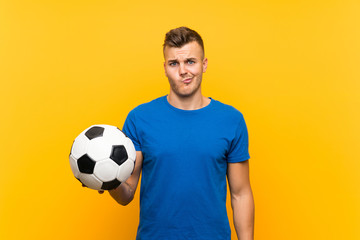 Young handsome blonde man holding a soccer ball over isolated yellow background with sad expression