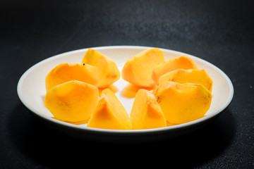 Persimmon fruit ripe small pieces in white plate top over black background.