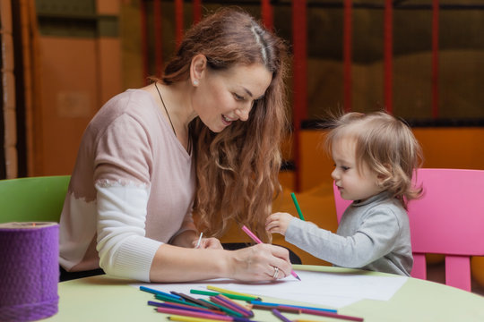 Nanny teaches a little girl how to draw with colored pencils while sitting at a table in a children's entertainment center
