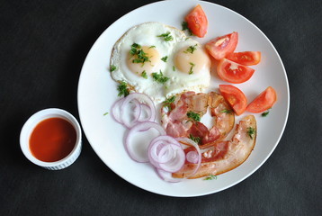 Fried eggs with tomatoes, red onion, bacon for breakfast on a white plate on the black background. Delicious homemade breakfast.