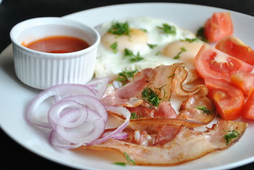 Fried eggs with tomatoes, red onion, bacon for breakfast on a white plate on the black background. Delicious homemade breakfast.