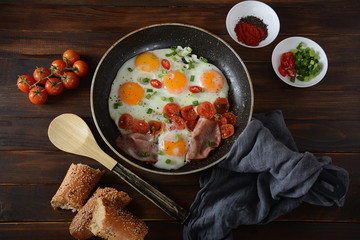 Fried eggs in a pan with cherry tomatoes, bacon and bread for breakfast on a wooden background. Delicious breakfast concept.