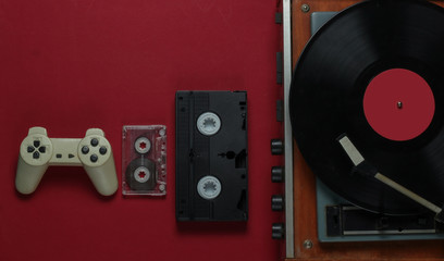 Flat lay retro media and entertainment. Vinyl record player with vinyl record, film camera, video cassette, audio cassette, gamepad on red background. 80s. Top view