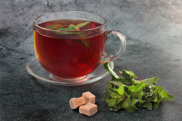 Abig glass mug of black tea with fresh mint leaves. More mint in the right corner. Three cubes of brown sugar. Dark gray background, back electric light