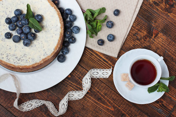 White plate with poppy cheesecake with blueberries. A plate cut from the left and above. A small cup of tea with two brown sugar in right bottom corner. All on a dark wooden table.