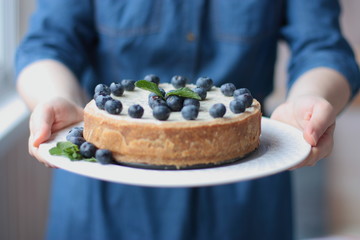 A woman in jeans-blue dress holds a home-made cheesecake with blueberries on a plate. Focus on the mint leaf. Day light.