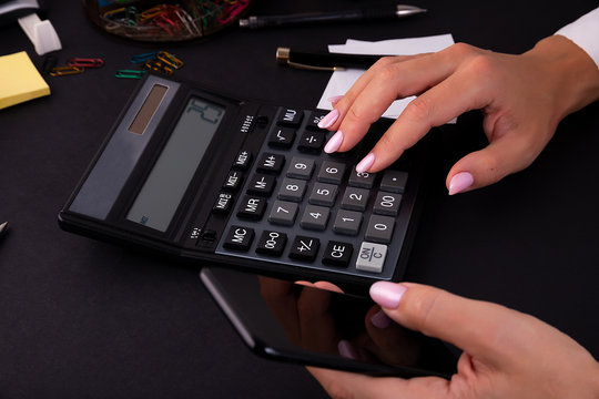 A woman presses a button on a calculator with her finger. She holds in her hand a black smartphone.
