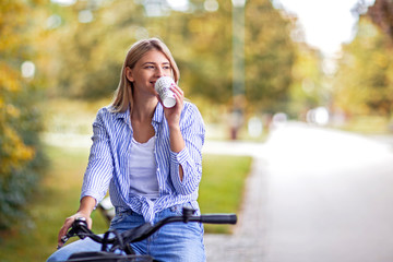 Fototapeta premium Beautiful young blonde girl on bicycle in park drinking coffee to go and smile