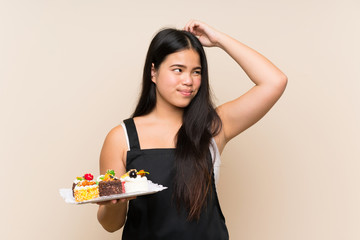 Young teenager Asian girl holding lots of different mini cakes over isolated background having doubts and with confuse face expression