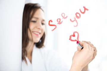 Hand of happy brunette girl writing and drawing heart with lipstick on mirror