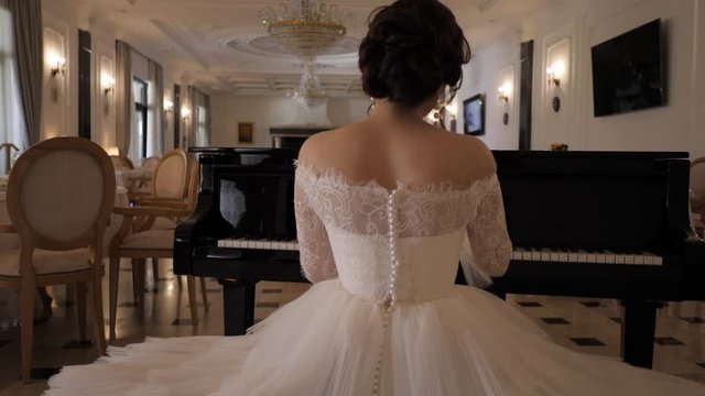 brunette lady in gorgeous dress with open shoulders decorated with pearls plays piano in wedding hall backside slow motion