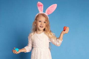 Portrait of a cheerful laughing girl in a white dress and pink rabbit ears holding Easter eggs in her hands. isolated on a blue background