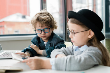 Serious schoolboy in eyeglasses looking at his clever classmate in hat