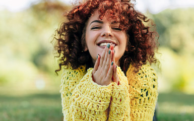 Portrait of cheerful young woman with curly hair smiling broadly with toothy smile. Female has positive expression, wearing yellow sweater and posing against nature background. People, lifestyle