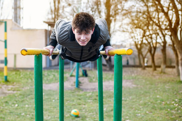 Obraz premium The guy doing sports on the playground near school and smiling, he is push up in horizontal bars on the playground