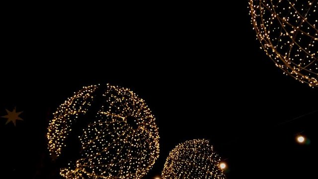 Walking underneath giant hanging christmas ornaments in a city, shaped like huge balls of light wrapped in fairy lights