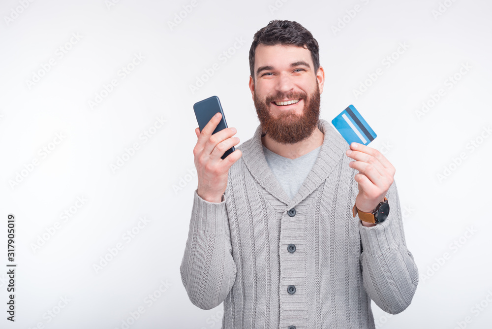 Wall mural handsome young bearded man is holding a credit card and a smartphone on a white background.