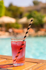 Red cocktail with ice with a straw on the background of the pool. vertical photo