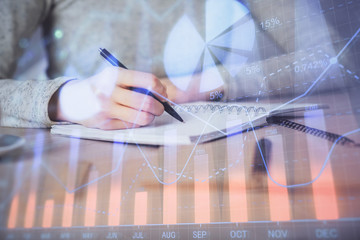 Hands writing information about stock market in notepad. Forex chart holograms in front. Concept of...