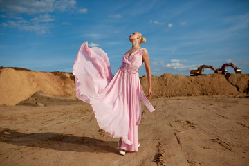 Fototapeta na wymiar Stylish female model with blonde hairstyle in flying pink dress, posing in a sand desert, at sunset background.