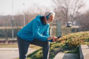 An elderly person wearing sports clothing using his phone and listening to music over headphones at...