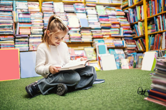 Adorable little Caucasian girl sitting on the floor in bookstore and reading stories. All around on shelves are books for kids.