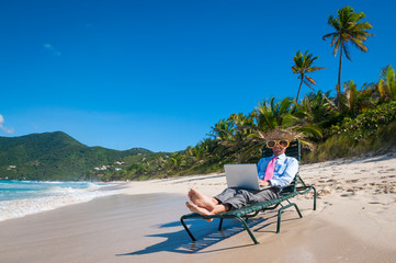 Businessman in straw hat and giant sunglasses typing on his laptop on a beach chair working remotely from the shore of an empty tropical beach