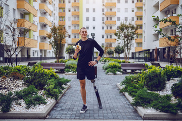 Full length of attractive caucasian sportsman with artificial leg standing in park surrounded by buildings and eating banana.