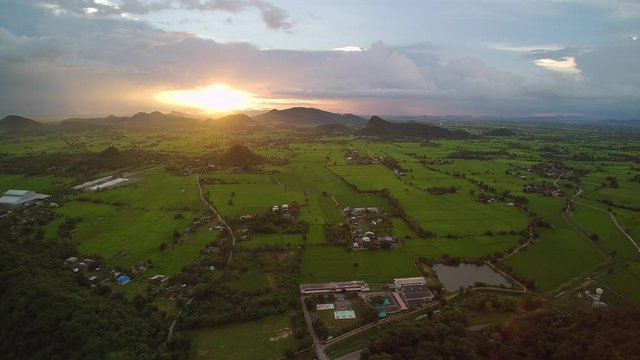 VDO. Aerial view evening above green rice paddy field, houses and buddhist temple with mountains and cloudy sky background, sunset at Wat Khao Chong Pran (Bat Temple), Photharam, Ratchaburi, Thailand.