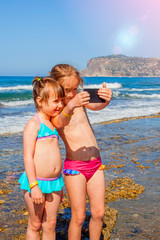 Fototapeta na wymiar Summer holiday, childhood and leisure concept. Two happy child girls taking selfie on smart phone on beach of sea. Vertical image.