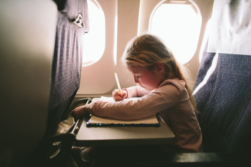 Little blonde girl sits on the plane and draws in notebooks during the flight