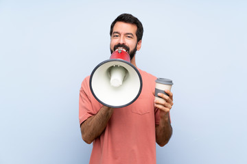 Young man with beard holding a take away coffee over isolated blue background shouting through a megaphone