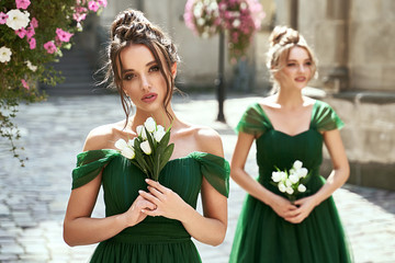 Two beautiful bridesmaids girls blonde and brunette ladies wearing elegant full length off-the-shoulder green chiffon bridesmaid dress and holding bouquets. European old town location for wedding day - obrazy, fototapety, plakaty