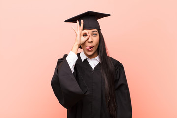 young latin woman student smiling happily with funny face, joking and looking through peephole,...
