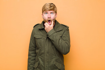 young blonde man with mouth and eyes wide open and hand on chin, feeling unpleasantly shocked,...