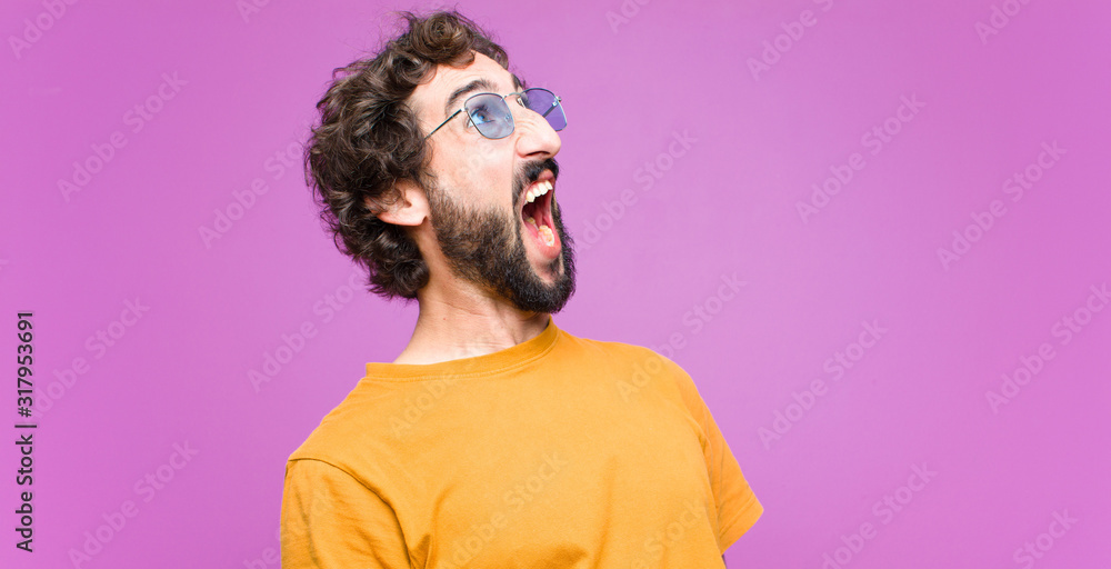 Wall mural young crazy cool man screaming furiously, shouting aggressively, looking stressed and angry against flat wall