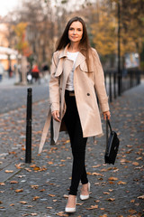 Outdoor photo of brunette lady walking on street background in autumn day.Fashion street style portrait. wearing dark casual trousers, white sweater and creamy coat.Fashion concept.