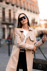 Obraz premium Beautiful brunette woman with phone walking on street.Fashion street style portrait. wearing dark casual trousers, white sweater,creamy coat and sunglasses .Fashion concept.Female business style.