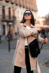 Beautiful brunette woman with phone walking on street.Fashion street style portrait. wearing dark casual trousers, white sweater,creamy coat and sunglasses .Fashion concept.Female business style.