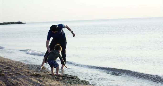 Father and son play on beach throwing rocks into ocean - wide slow motion shot