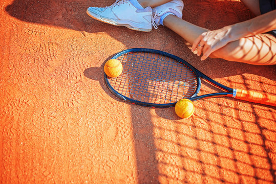Legs and racket of female player that seets on the ground of the court at daytime.