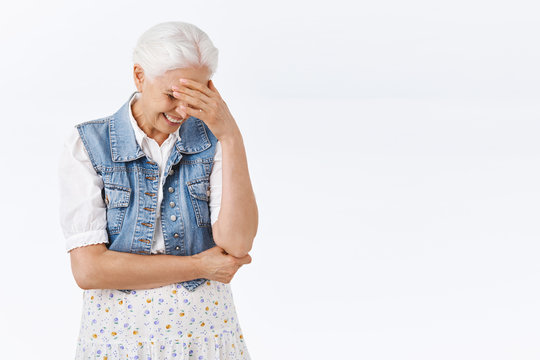 Carefree, amused charming modern granny with grey combed hair in stylish denim vest, dress, laughing over hilarious kid outfit or joke, look down chuckling and smiling, face palm, touch forehead