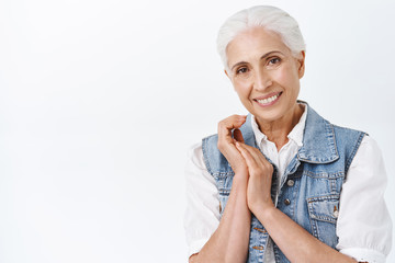 Tenderness, aging and cosmetology concept. Modern cute and happy smiling senior woman with combed grey hair, clasping hands and gazing camera delighted, taking care of skin