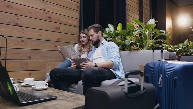 Front view of likable romantic joyful couple which sitting together in hotel lobby and using tourist map and tablet pc to search interesting places to visit