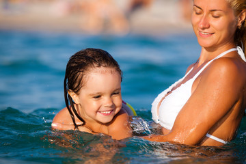 Young blond mother in blue bikini standing in water and helping to swim her small smiling daughter in sunshine on summer day. Family vacations and travelling concept