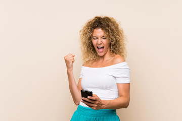 Fototapeta premium Young blonde woman with curly hair over isolated background with phone in victory position