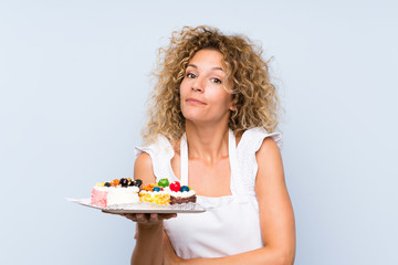 Young blonde woman with curly hair holding lots of different mini cakes making doubts gesture while lifting the shoulders