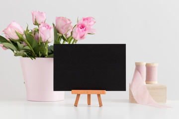 Black card mockup with silk ribbons and pink roses in a pot on a white table.